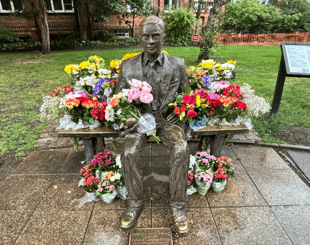 Life sized statue of a a man sitting on a park bench. The bench is covered in and surrounded by flowers